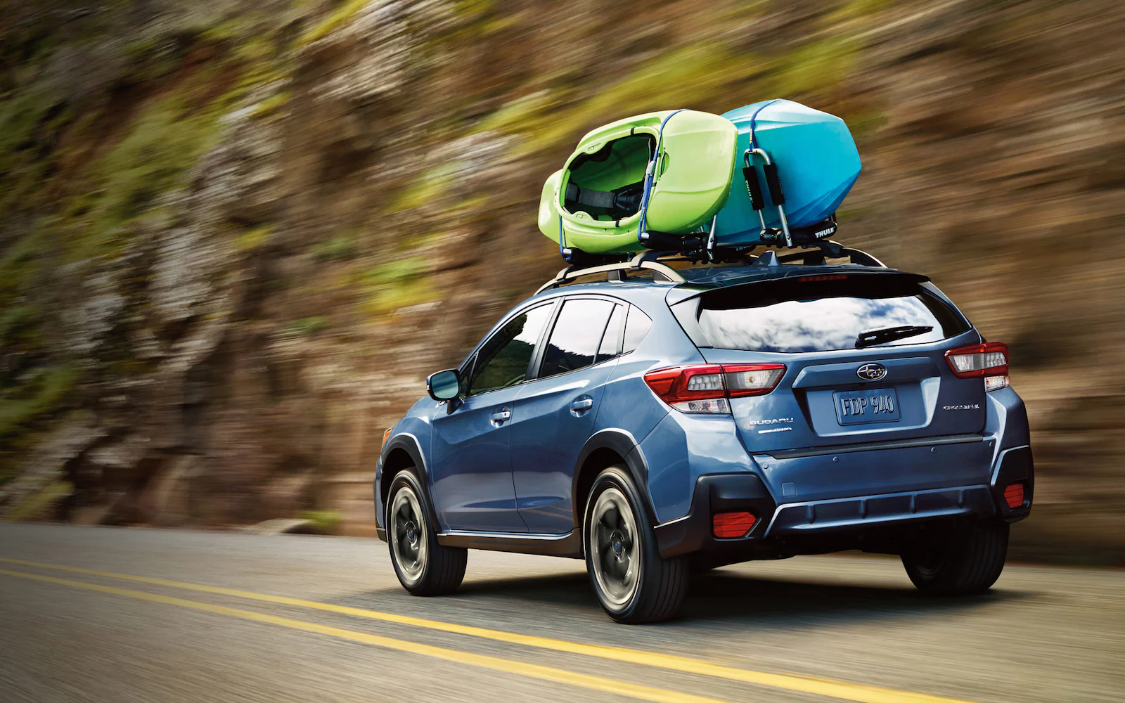 A blue Subaru Crosstrek with two kayaks on roof driving on a mountain road